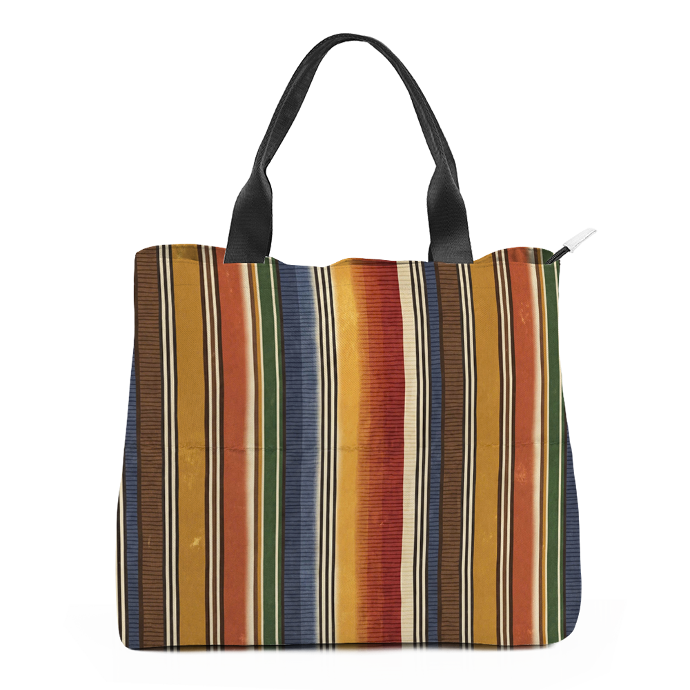 Sedona tote bag turned slightly so the side panel shows, printed with vertical serape stripes in mustard, burnt rust, brick red, olive green and denim blue split by tight cream hairlines; flat black webbing handles arch above the opening and a pale zip pull hangs at the top right corner.