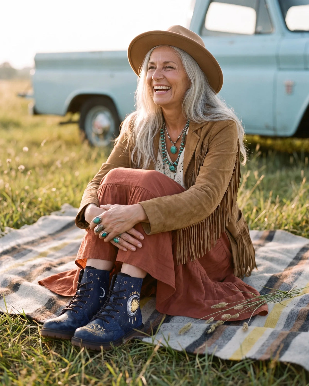 A woman with long silver hair in a felt hat and fringed jacket sits on a striped blanket in a meadow, wearing navy Celeste boots with a gold sun and crescent moon