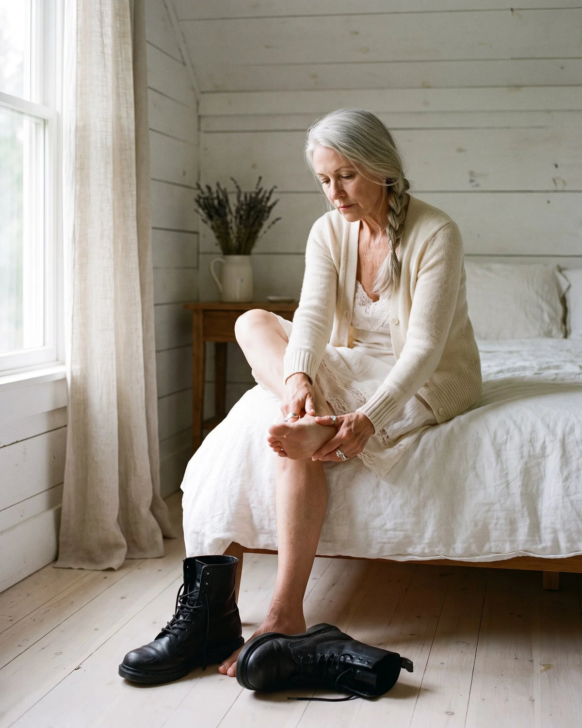 An elegant woman with a long silver braid sits on her bed in a white farmhouse bedroom rubbing her bare foot, plain black combat boots kicked off on the floor