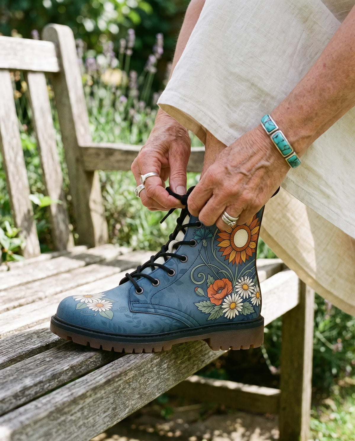Hands with silver rings and a turquoise bracelet pull the laces of a blue Daisy boot snug on a garden bench