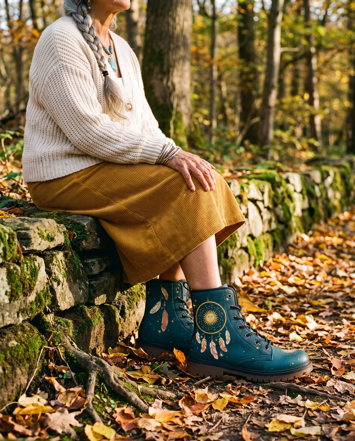 A woman with a long grey braid sits on a mossy stone wall in autumn woods, wearing teal Willow boots with a gold dreamcatcher